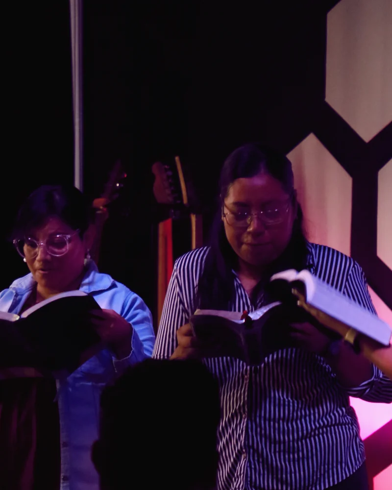Dos mujeres leen y cantan con su Biblia abierta frente a la pared hexagonal iluminada